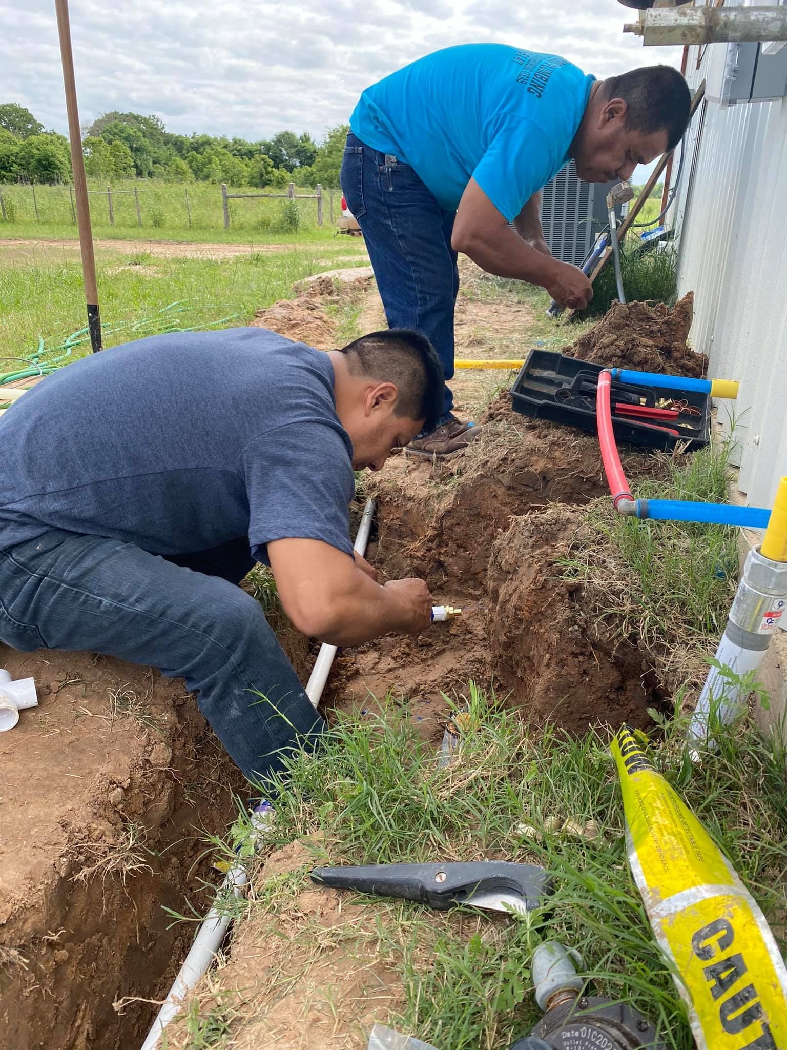 Two workers installing pipes in a trench near a building. They use tools, wearing casual clothes, outdoors.
