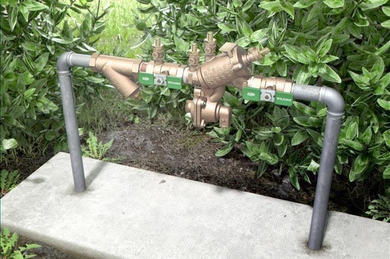A backflow preventer assembly with pipes, valves, and a concrete base, nestled in green foliage.