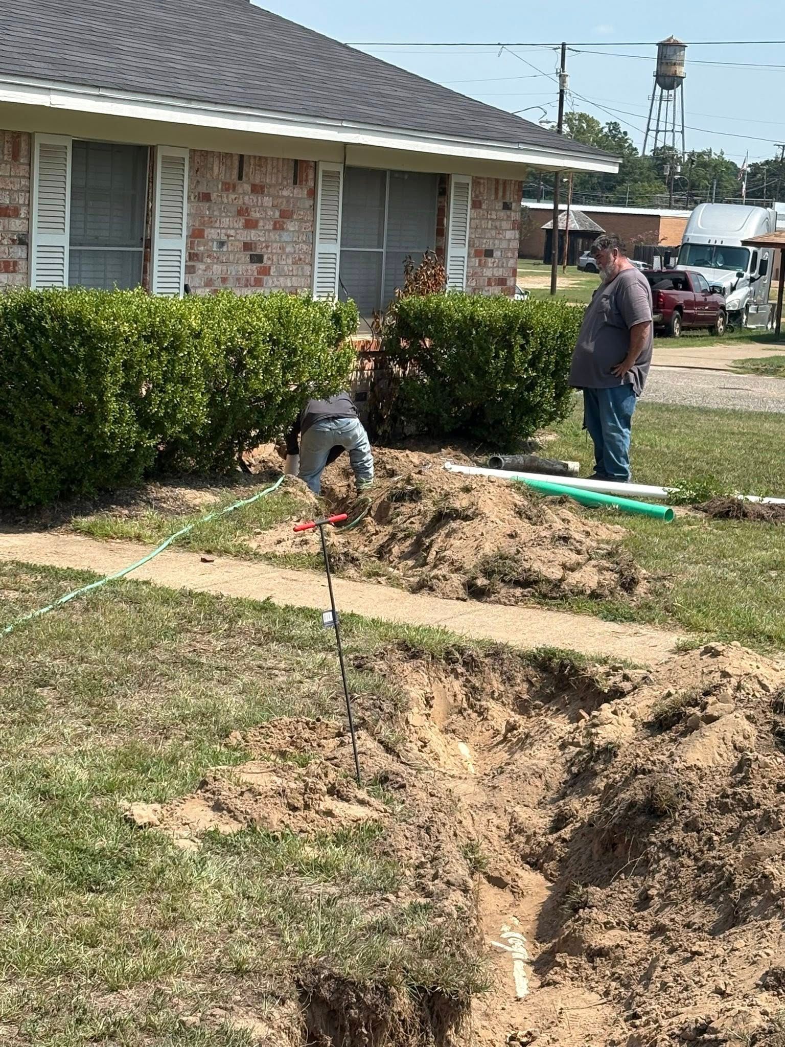 Workers digging in yard near a house. One is in the trench; another stands near.