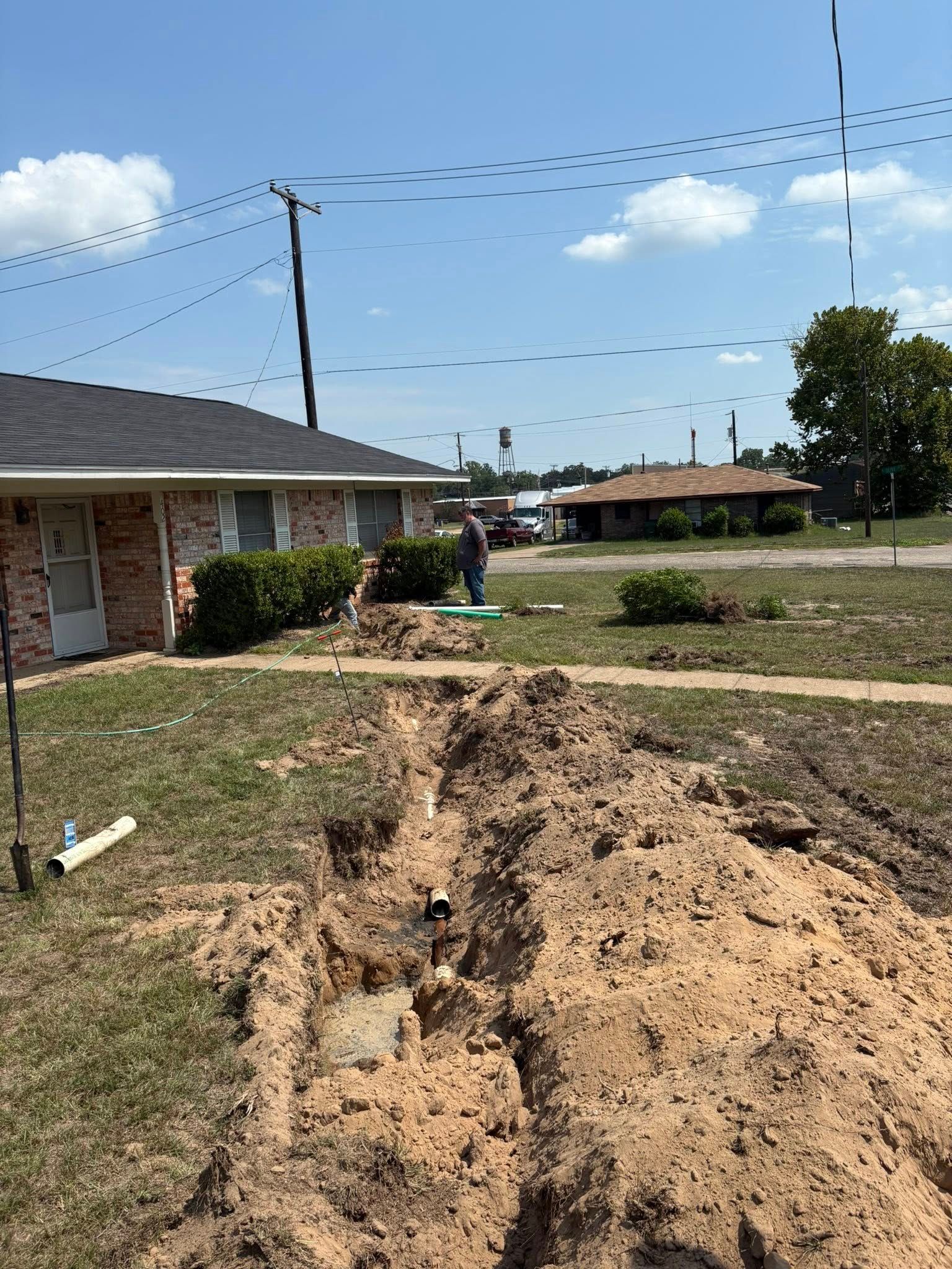 Dug trench in front yard of brick house; person standing nearby. Sky with utility lines overhead.