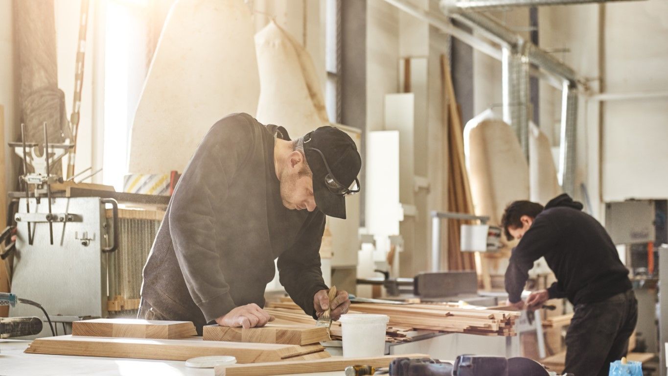 Two people working at a woodworking bench in a bright workshop, sanding wood pieces.