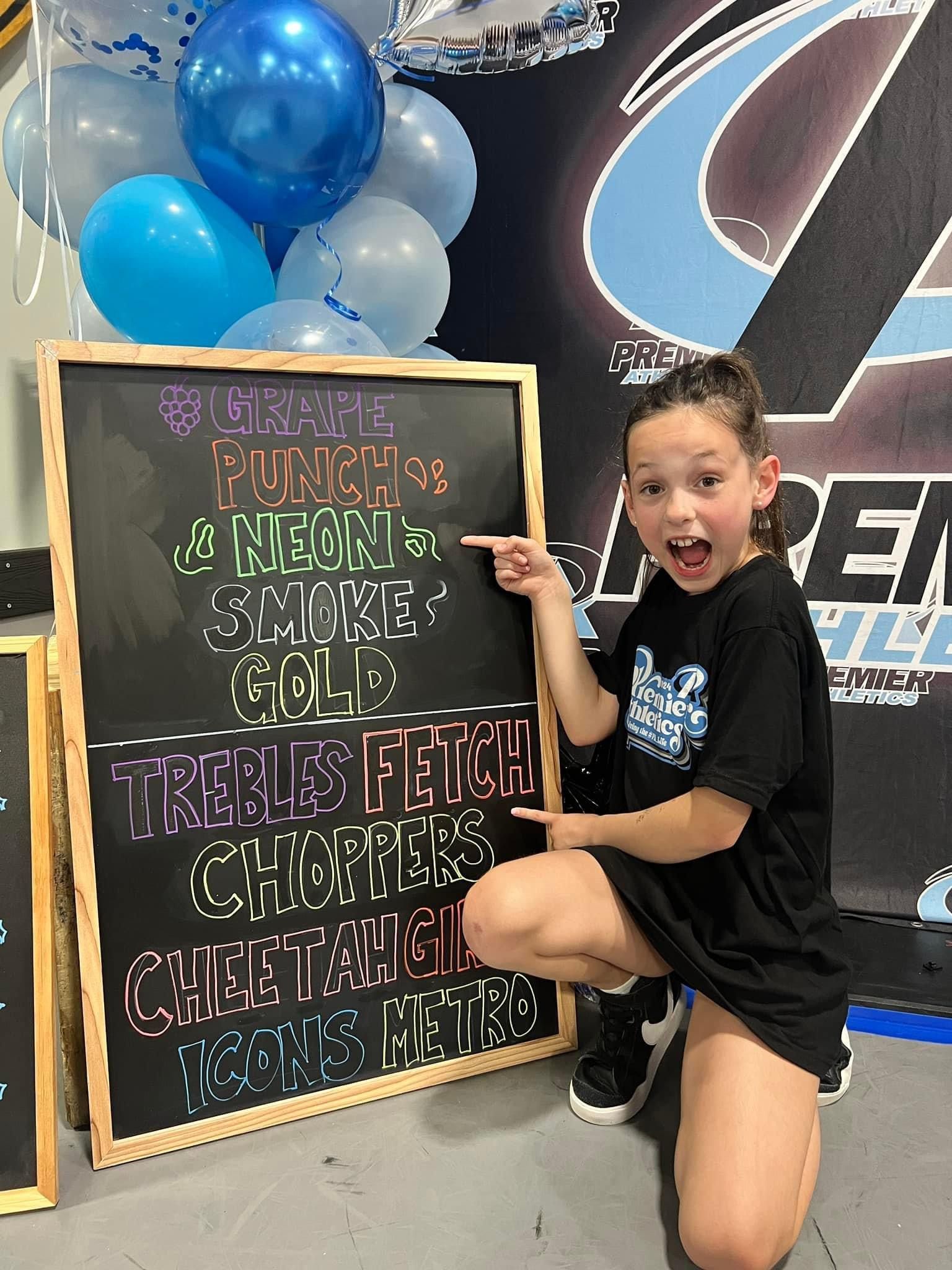 A little girl is kneeling down next to a chalkboard with balloons in the background.