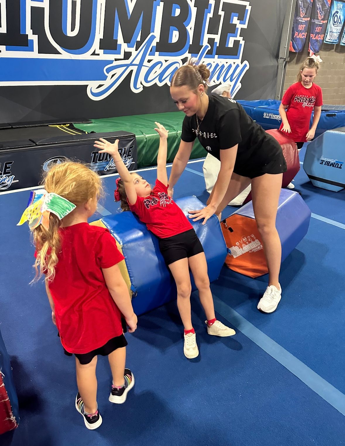 A woman is helping two young girls do exercises on a gym floor.