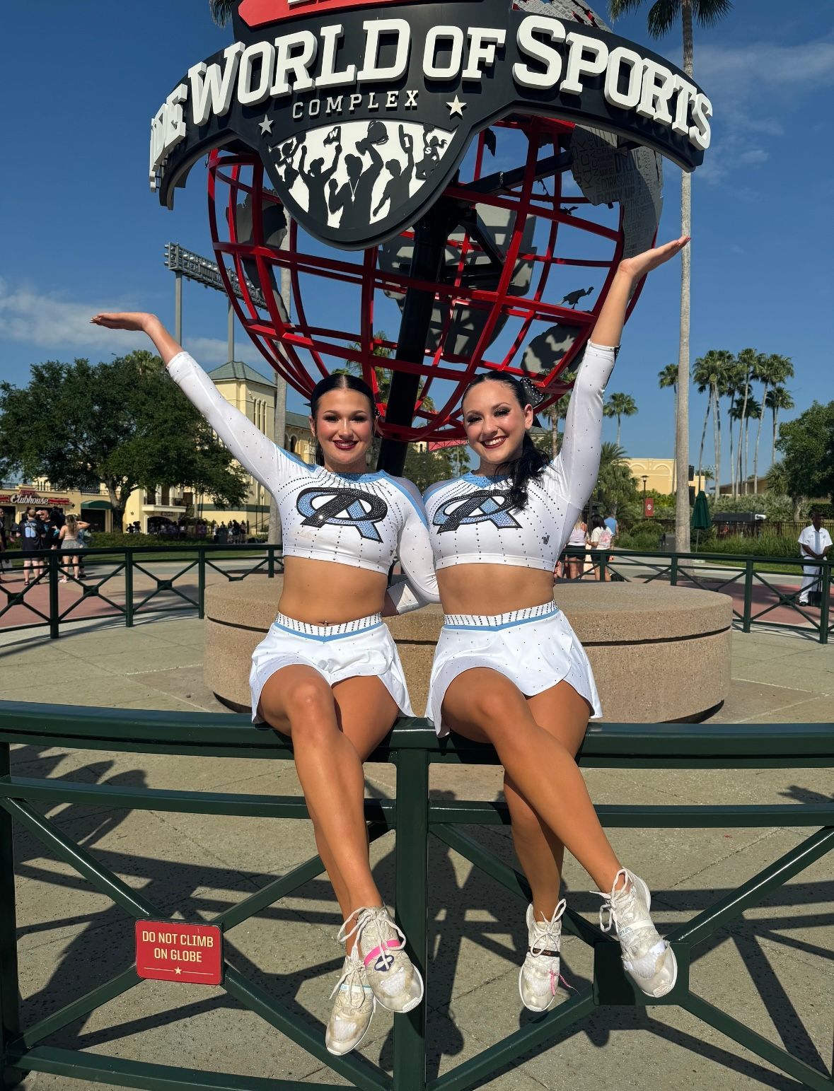 Two cheerleaders are sitting on a fence in front of a world of sports sign.