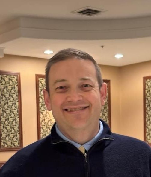 Man smiling, wearing a blue sweater and light blue shirt, in a room with beige walls and artwork.