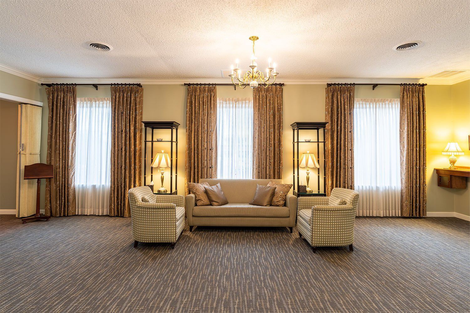 Empty, carpeted room with sofa, chairs, and windows with curtains; chandelier overhead.
