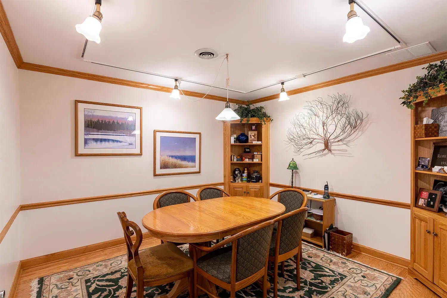 Dining room with oval table, chairs, artwork, bookshelves, and overhead lighting.