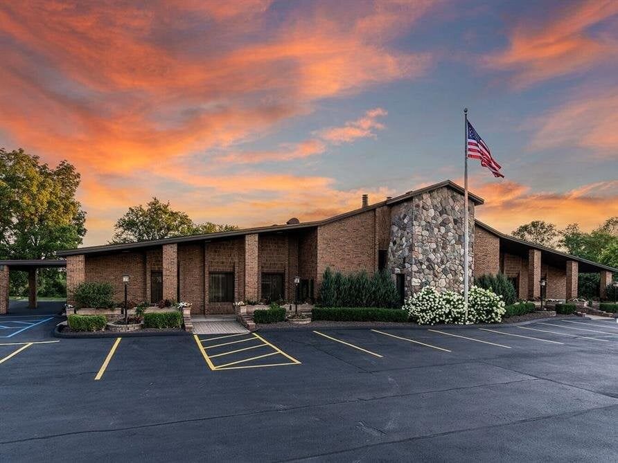 Exterior of a brick building with stone facade, American flag, and dark parking lot under a sunset sky.