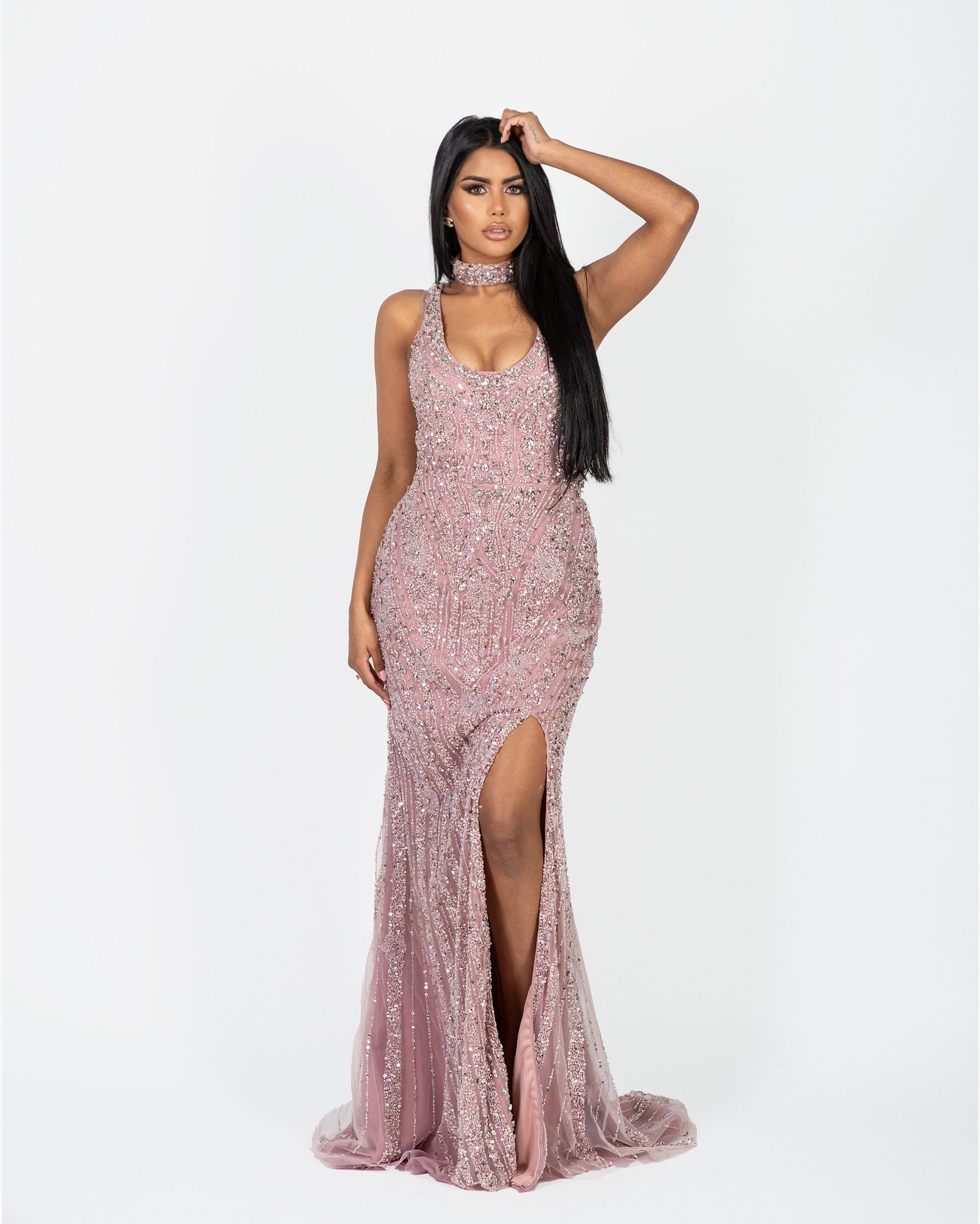 Vrouw in een glinsterende, roze jurk met een hoge split en ontroerend haar, poserend tegen een witte achtergrond.