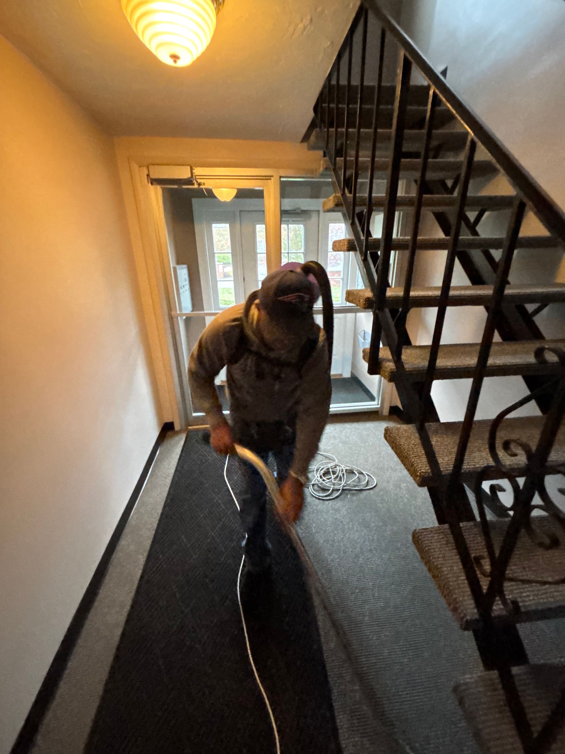 Person vacuuming a dark gray carpeted hallway with stairs on the right. A door is at the end.