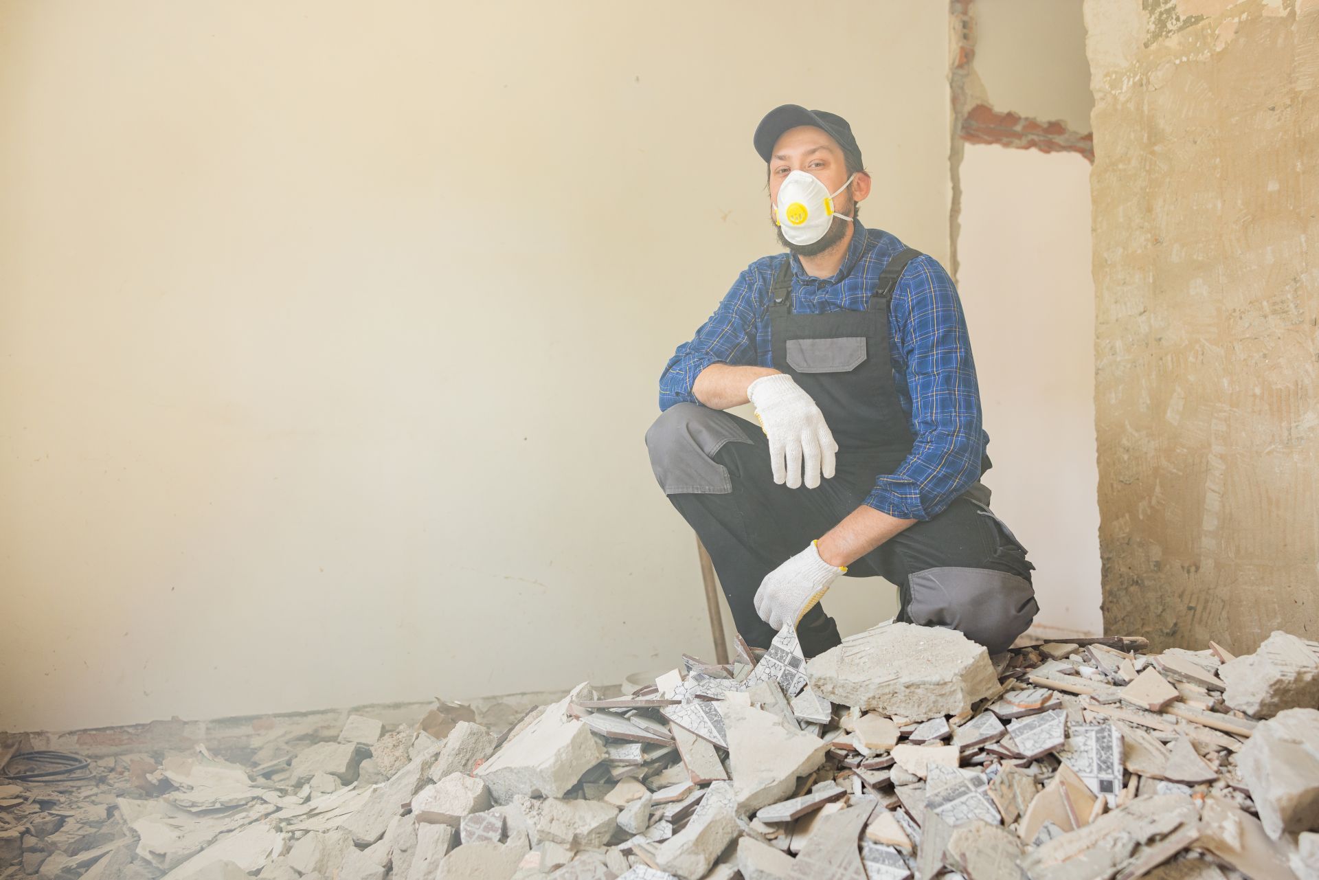 Construction worker squats amid debris, wearing a respirator, cap, and gloves. Inside a room with damaged walls.