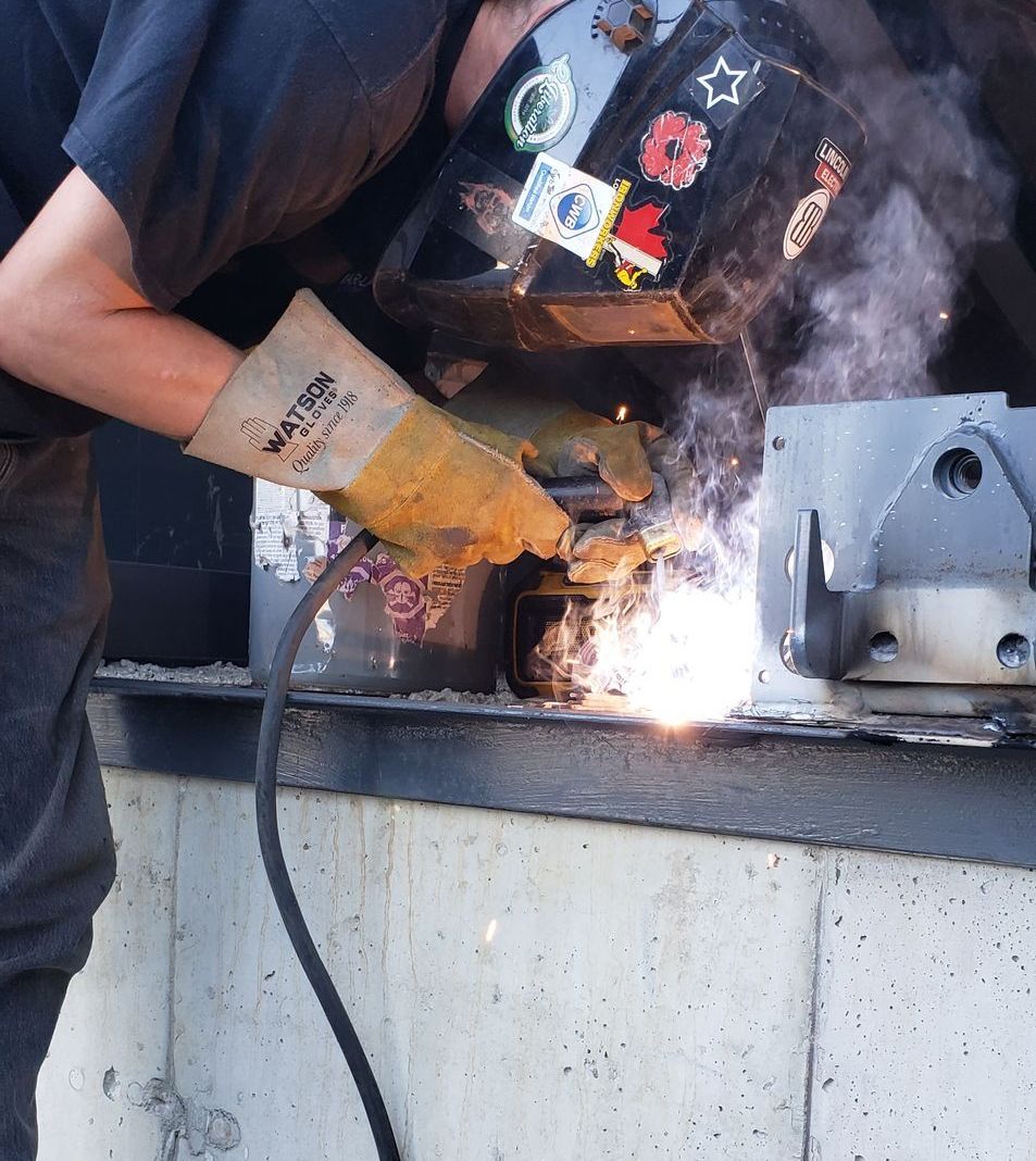 A person in a welding mask and gloves welds metal, producing a bright light and smoke near a concrete wall.