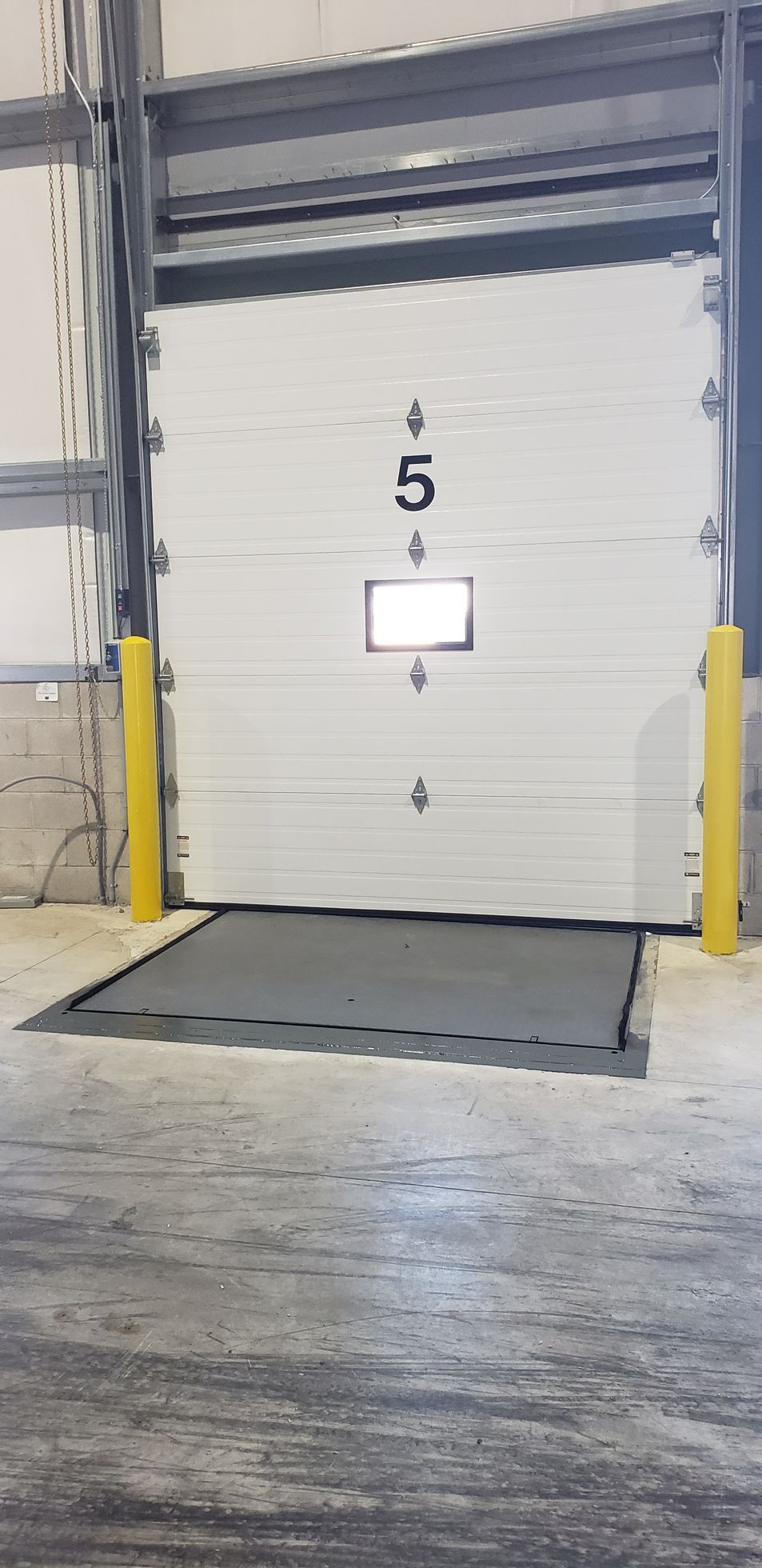 A loading dock bay door. The door is white and has a loading platform in front of it, 2 yellow bollards on either side.