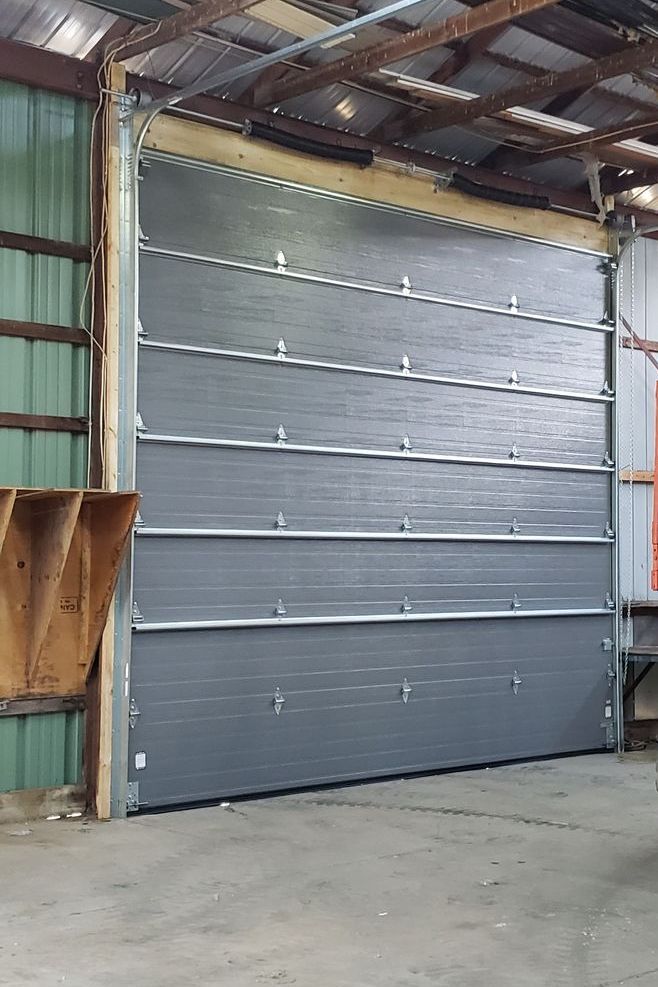 A closed, gray overhead garage door in a barn. The door is framed in wood, with a corrugated metal roof overhead.