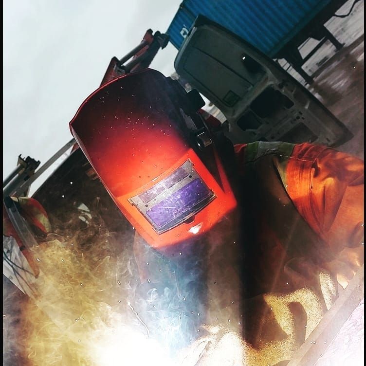 Welder in a red helmet, welding with sparks, with a truck and container in the background.