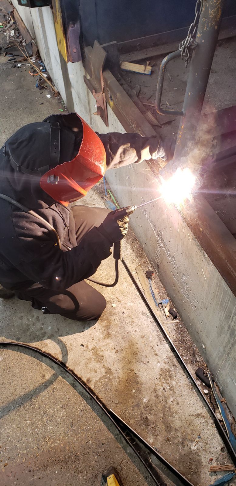 A person welding metal, wearing a red welding helmet. The action is taking place outdoors, and there is a lot of light.