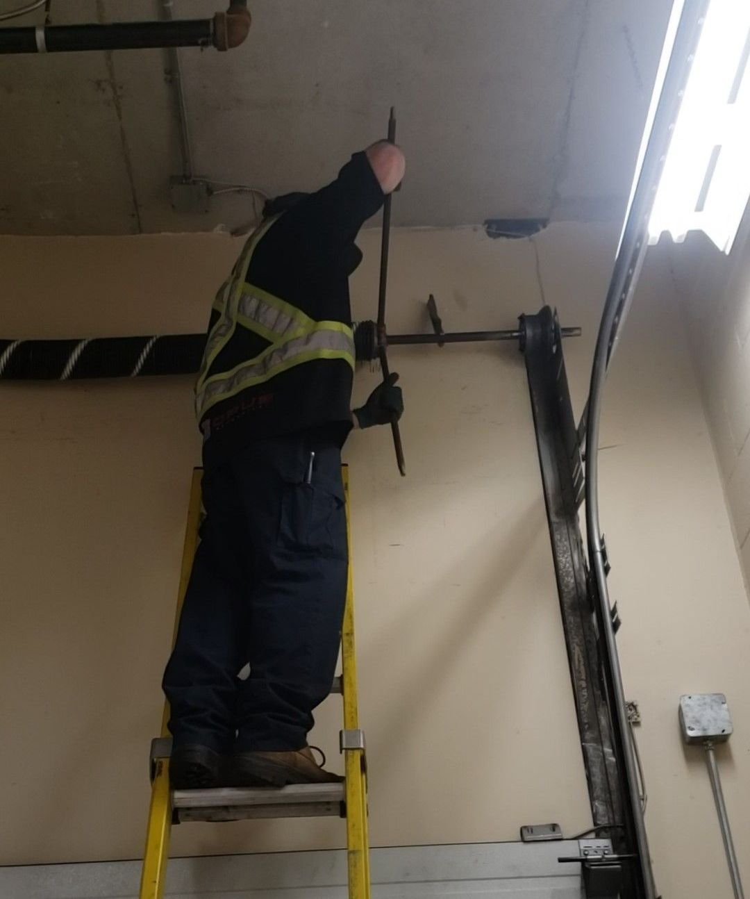 A person in a safety vest on a ladder, working on a garage door mechanism. The ceiling and wall are visible.