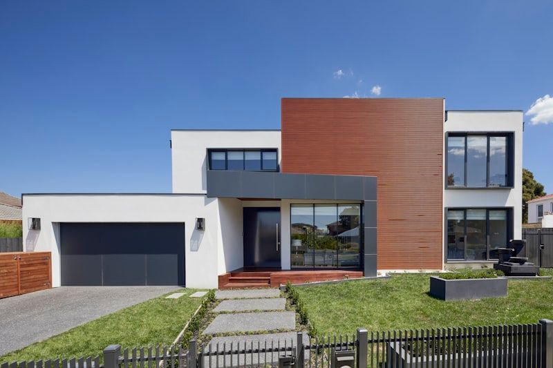 Modern two-story house with white, wood, and glass facade, garage, and fenced front lawn