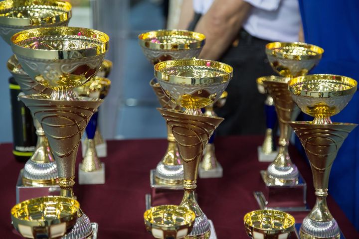 Diversi trofei d'oro e d'argento disposti su una superficie rosso scuro.