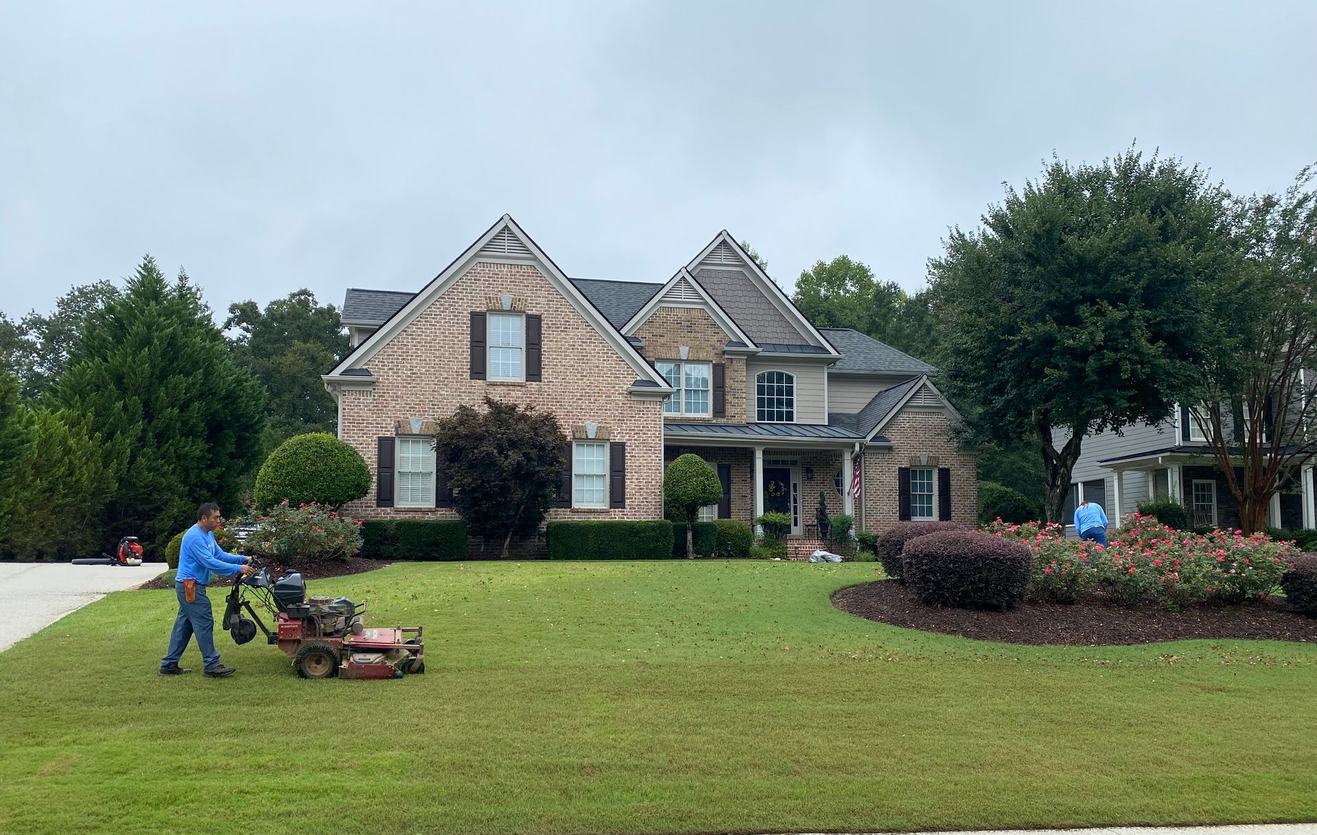 A man is mowing the lawn in front of a large brick house.