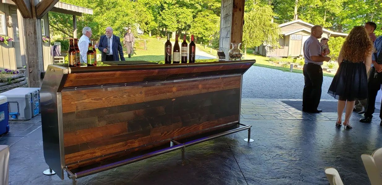 A group of people are standing around a wooden bar with wine bottles on it.