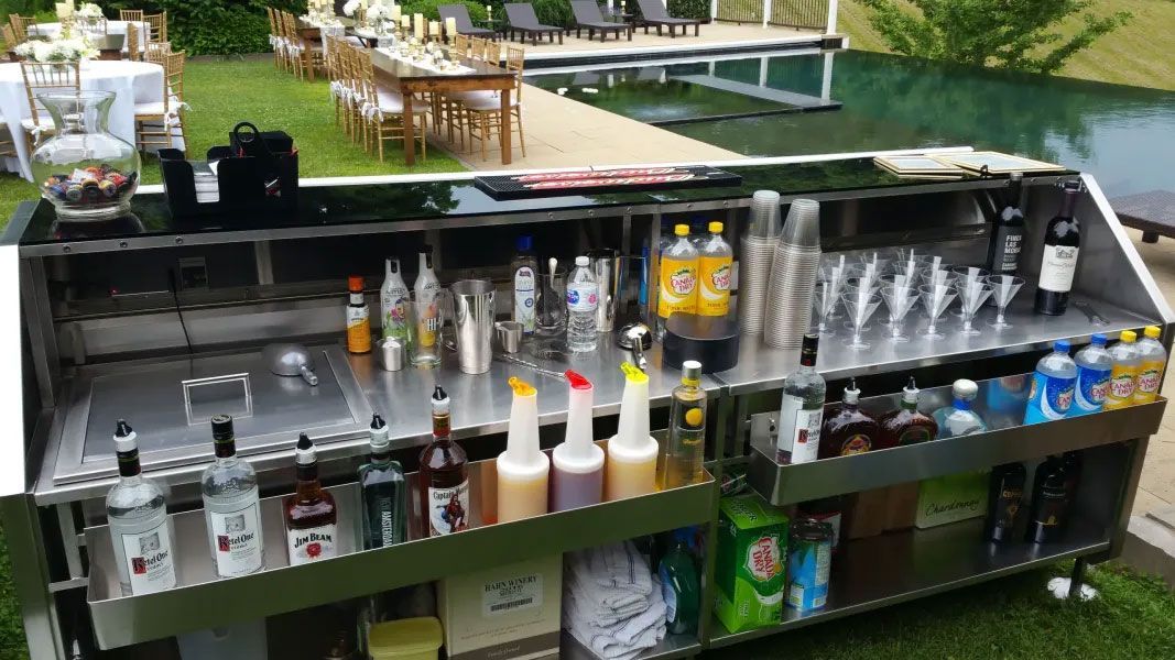 A stainless steel bar filled with bottles and glasses is sitting in the grass in front of a pool.