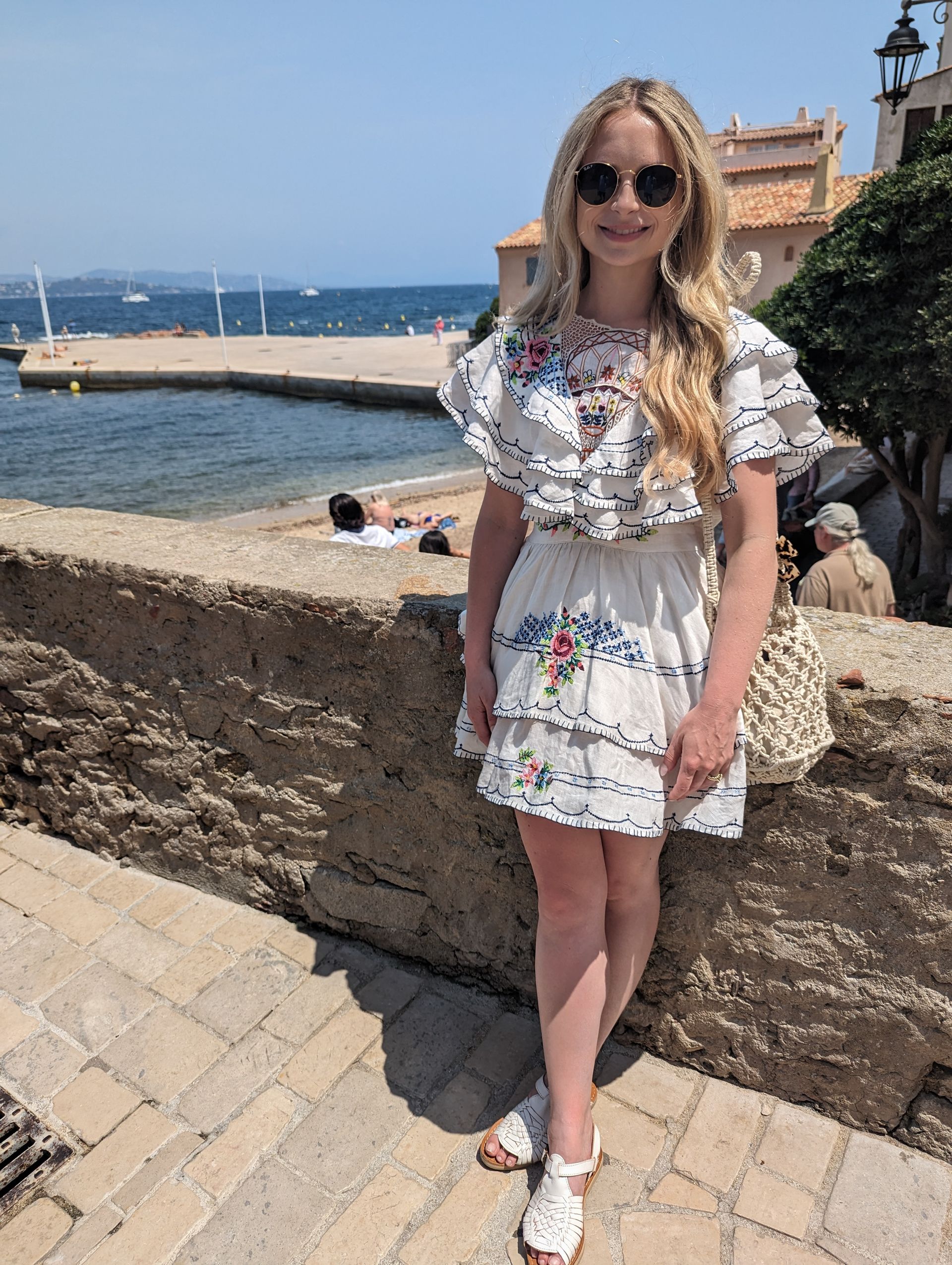 Woman in embroidered dress and sunglasses stands by a stone wall overlooking the sea.