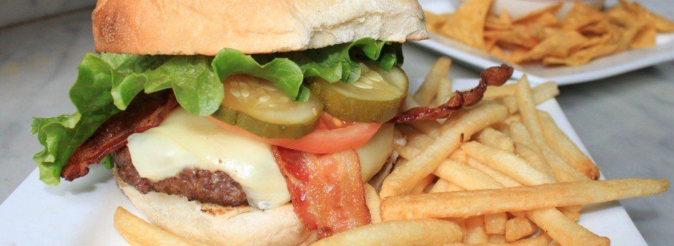 A cheeseburger with bacon, lettuce, tomato, and pickles, served with a side of french fries on a white plate.