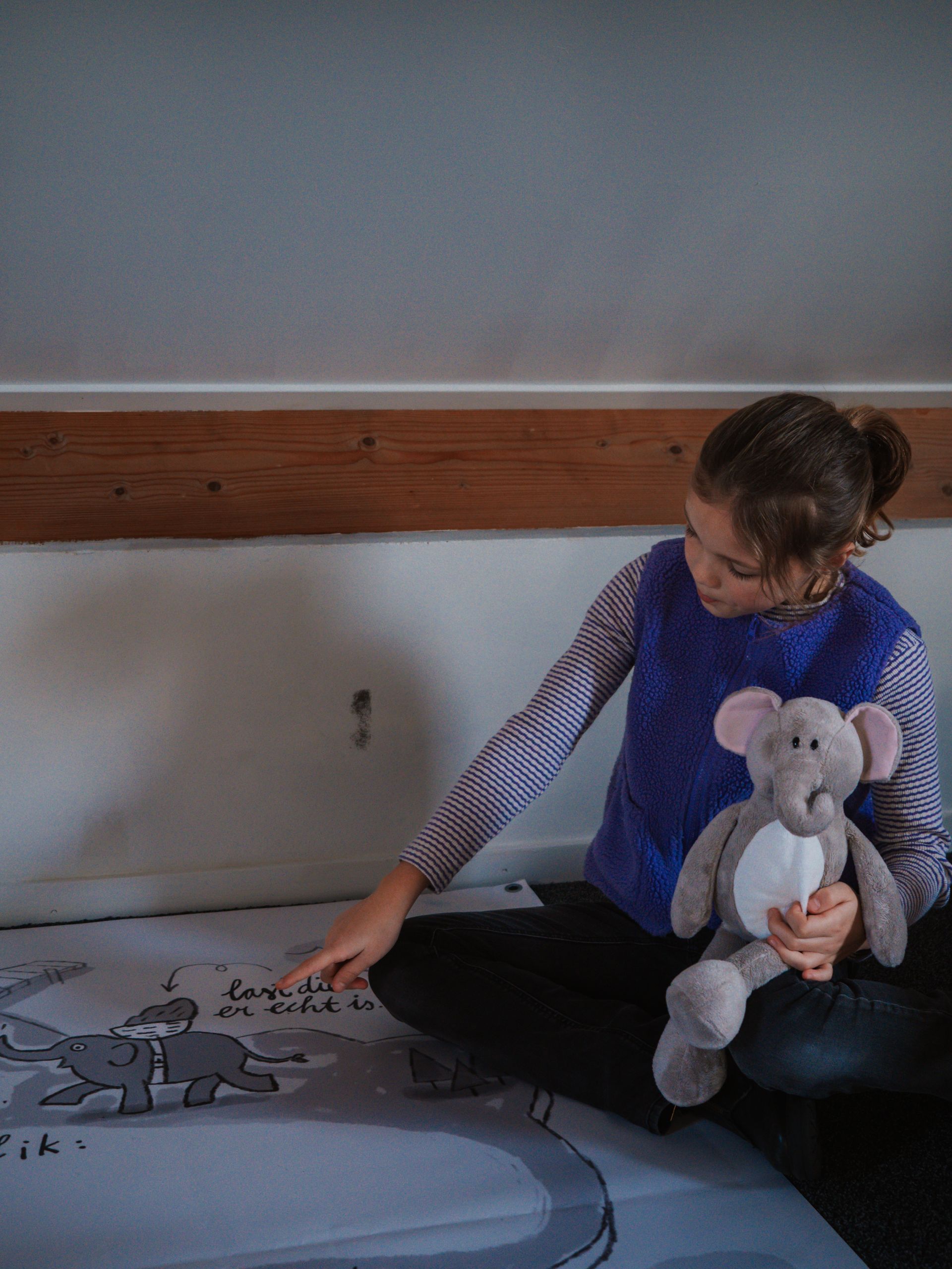 Een klein meisje zit op een bed met een knuffelolifant in haar armen.