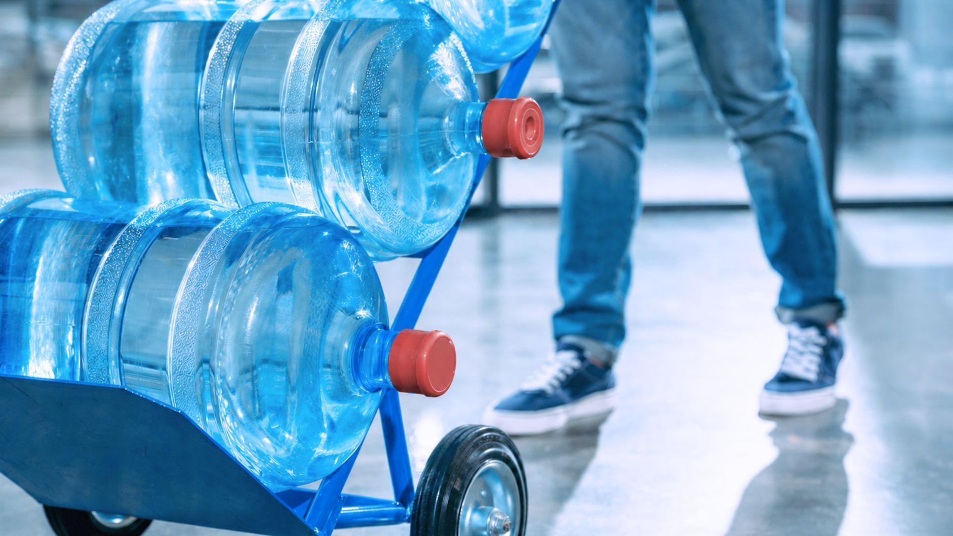 Stacked blue water jugs on a hand truck beside a person walking indoors delivering fresh spring water - Indian Peaks Spring Water