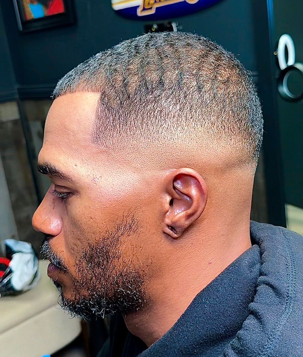 Man with faded haircut and groomed beard, side profile view. Dark hair, light skin, wearing a dark hoodie.