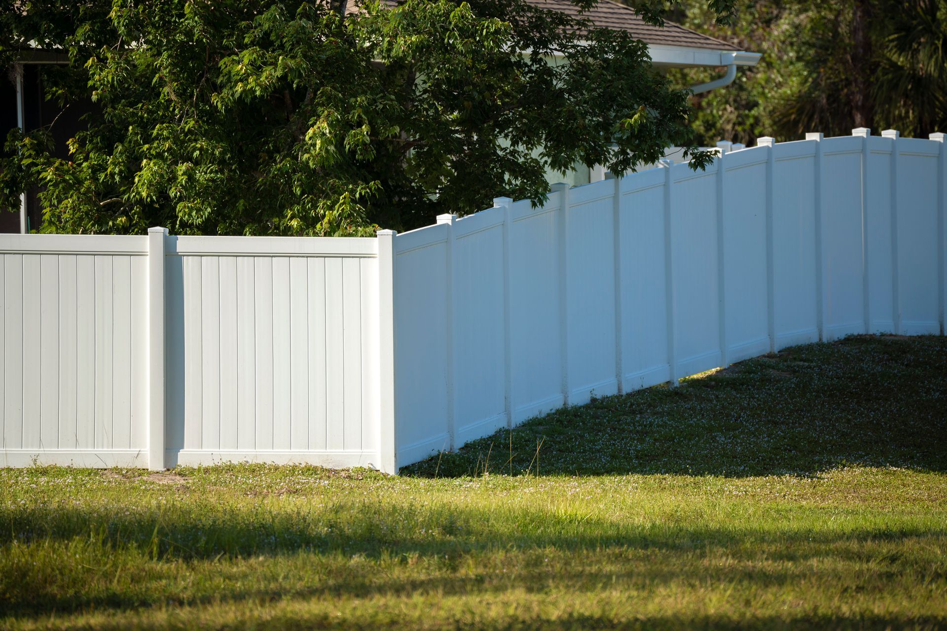White vinyl fence along grassy yard, with trees and a house in the background.