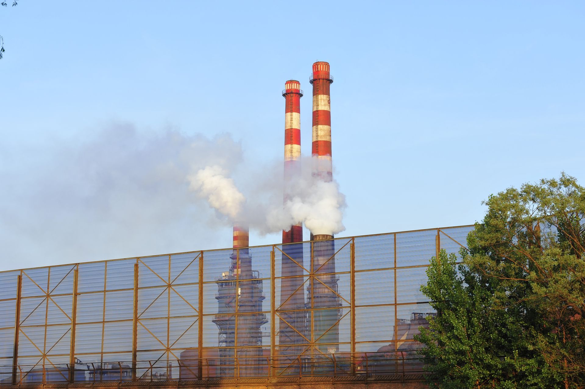 Smokestacks emitting smoke into the sky, behind a metal fence and trees.