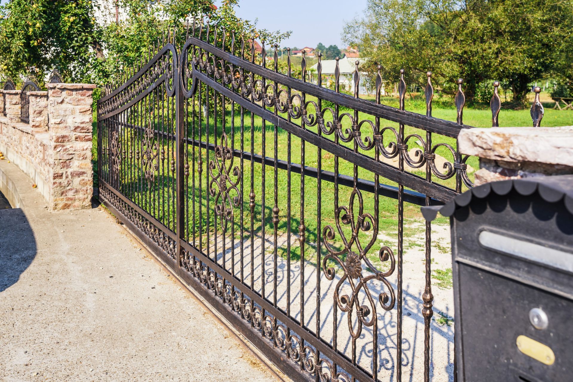 Black wrought-iron driveway gate, brick pillars, and mailbox. Green lawn and trees in the background.