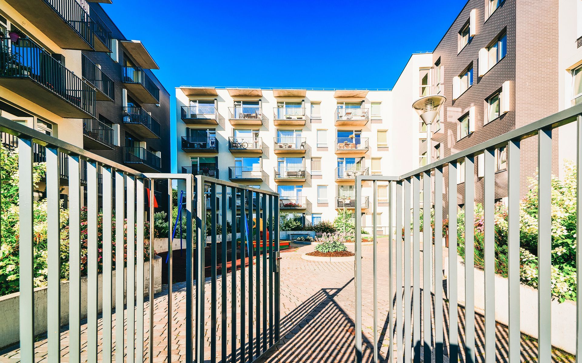 Apartment complex courtyard with metal gate, brick path, and multiple stories of balconies under a blue sky.