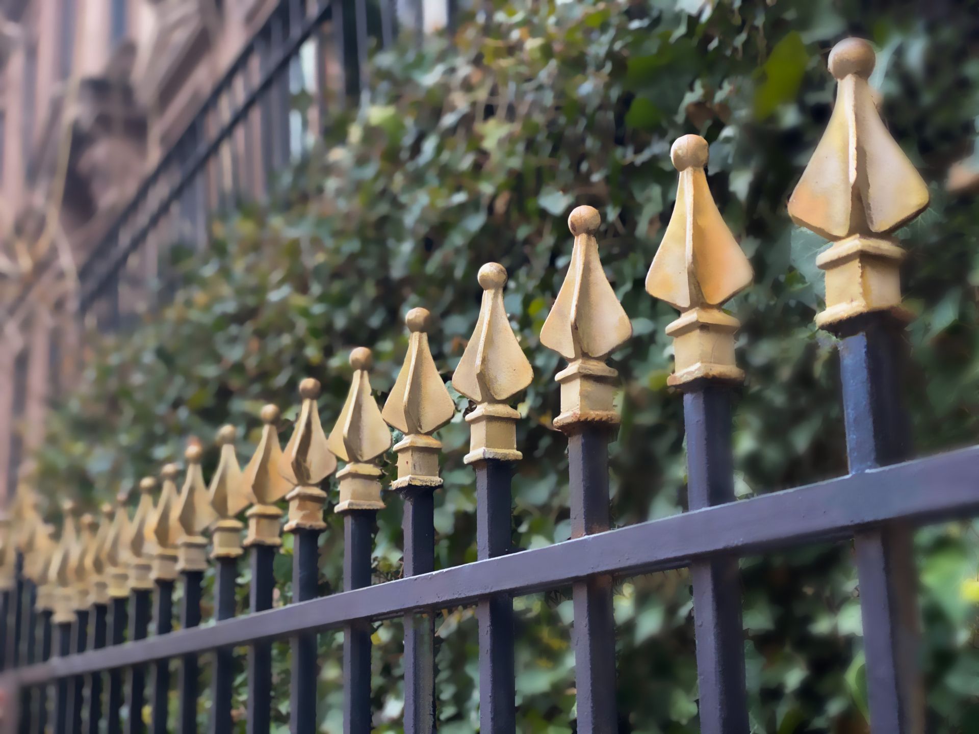 Black iron fence with gold-tipped decorative points, in front of greenery and a brick building.