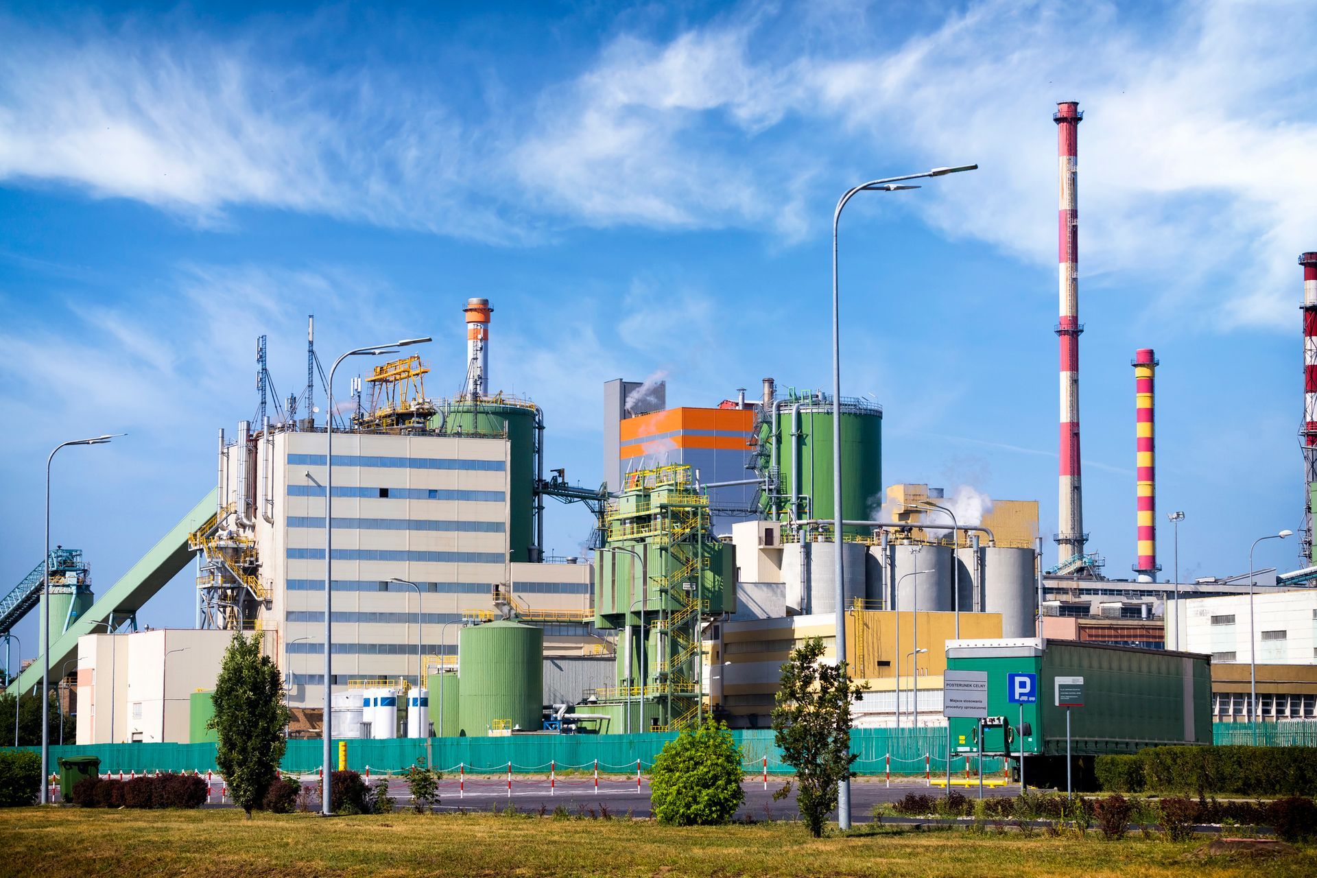 Industrial plant with several smokestacks, tanks, and buildings against a blue sky with clouds.