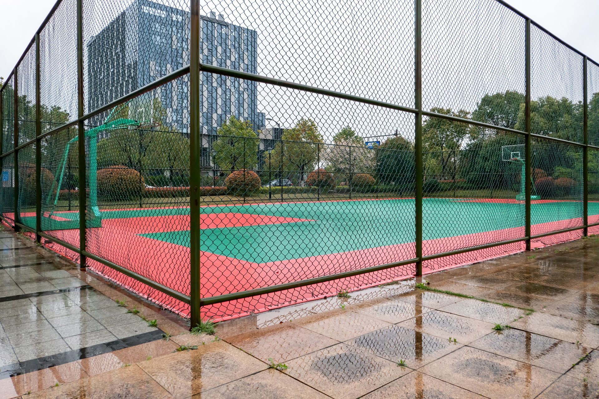 Basketball court with green and red surface, enclosed by a fence, set in a city park.