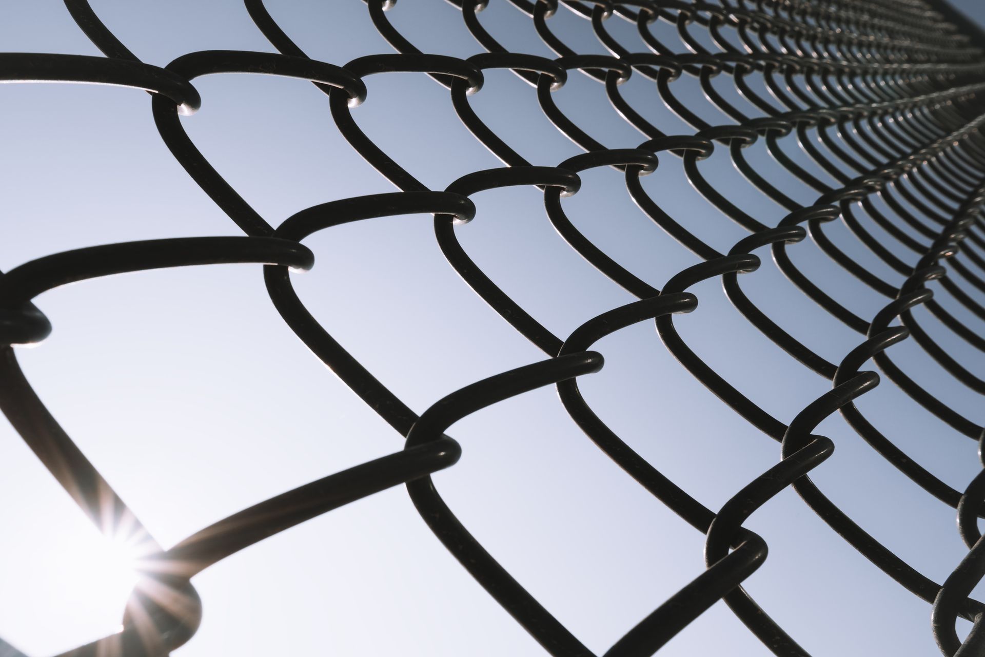 Chain-link fence against a bright sky, sunlight shining through.