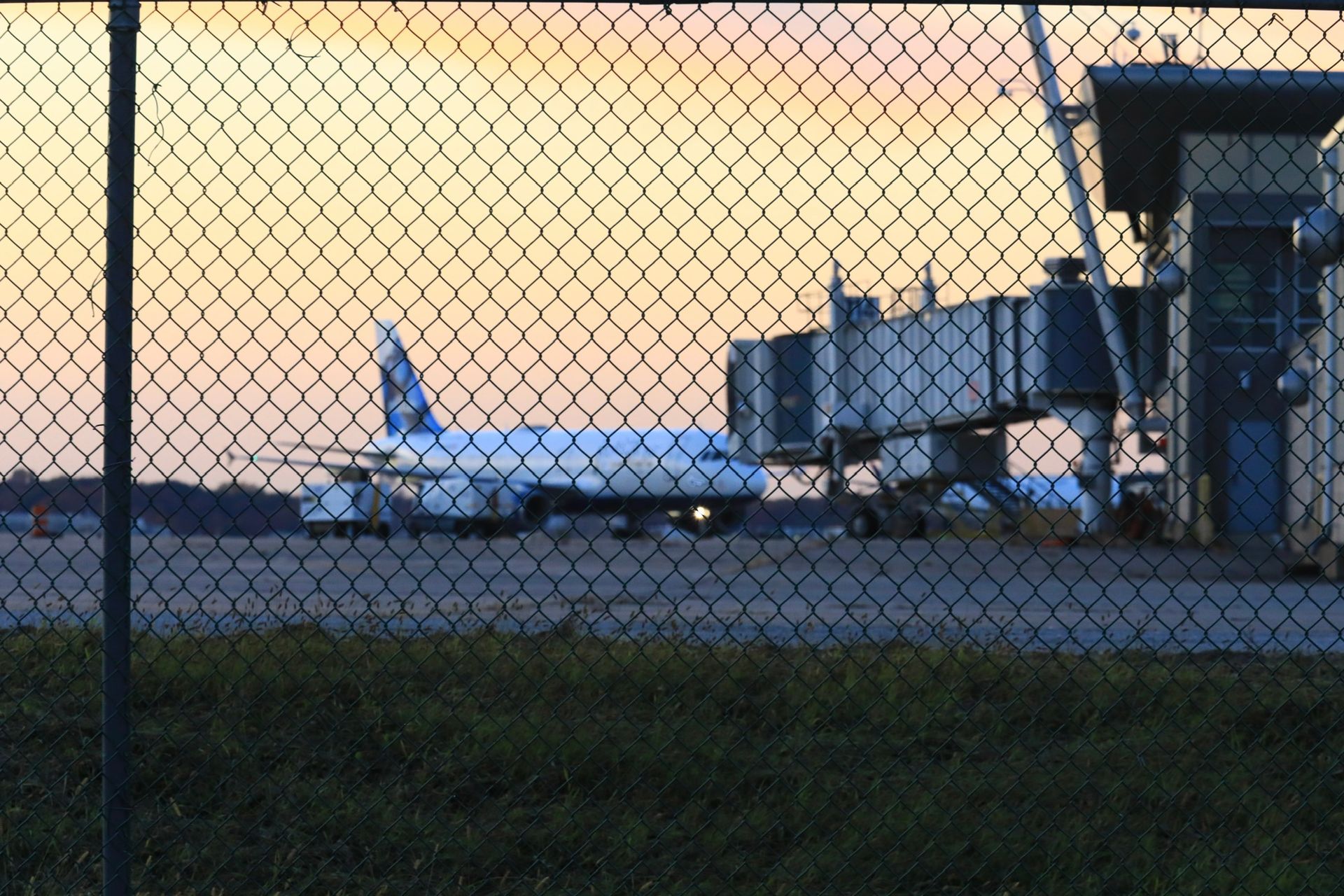 Airplane on tarmac near a jet bridge, seen through a chain-link fence at dusk.