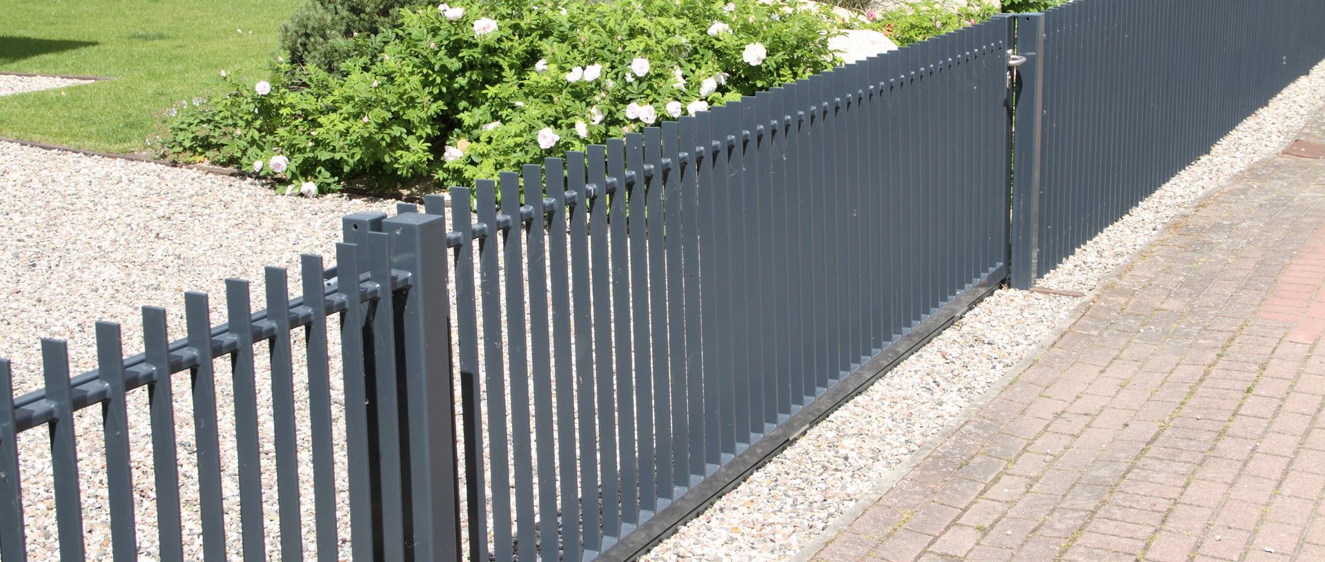 Gray metal fence along a walkway, with grass and bushes in the background.