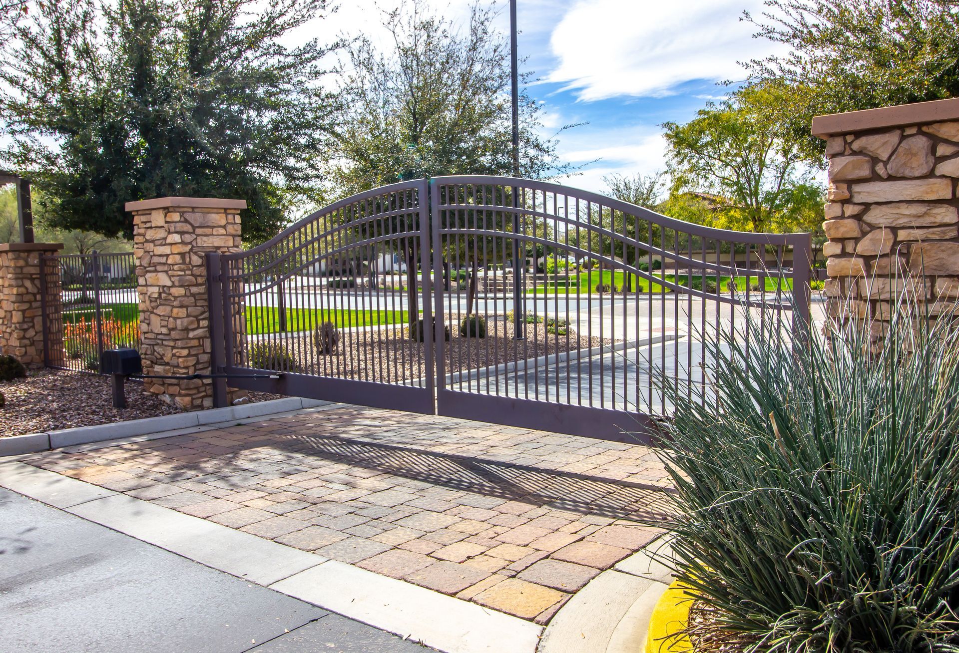 Ornate, wrought-iron entrance gates with brick columns. Paved entryway with landscaping under a blue sky.