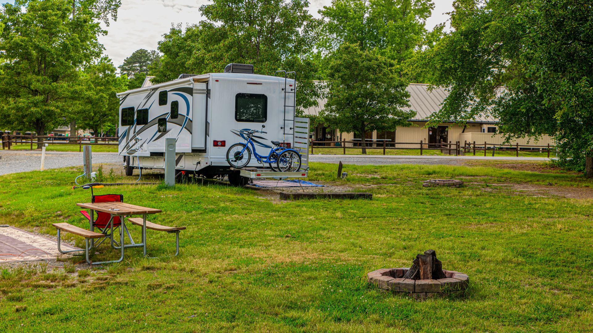 RV parked on grassy campsite, picnic table, fire pit, trees and buildings in the background.