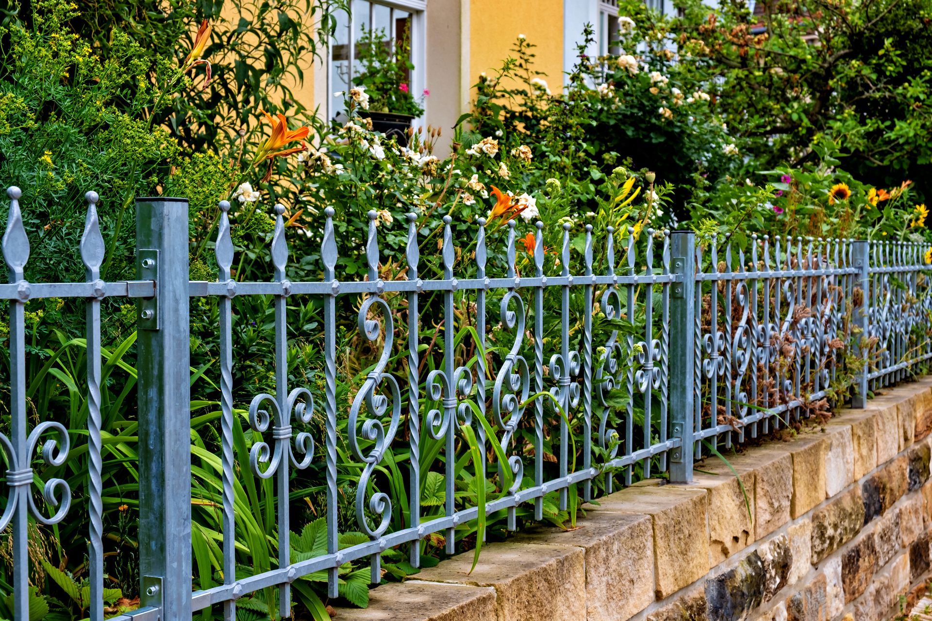 Ornate metal fence atop a brick wall, with greenery and building in the background.