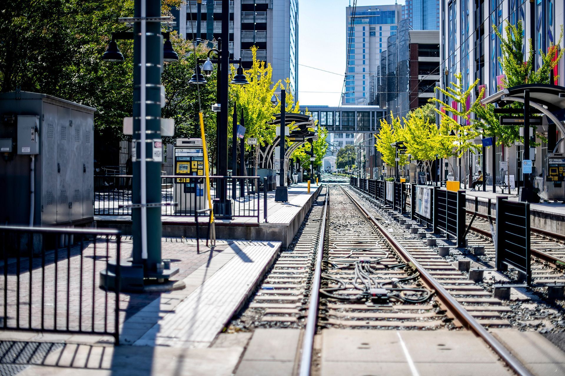 Train tracks at a city station. Buildings line the tracks. Green trees and a sky bridge are visible.