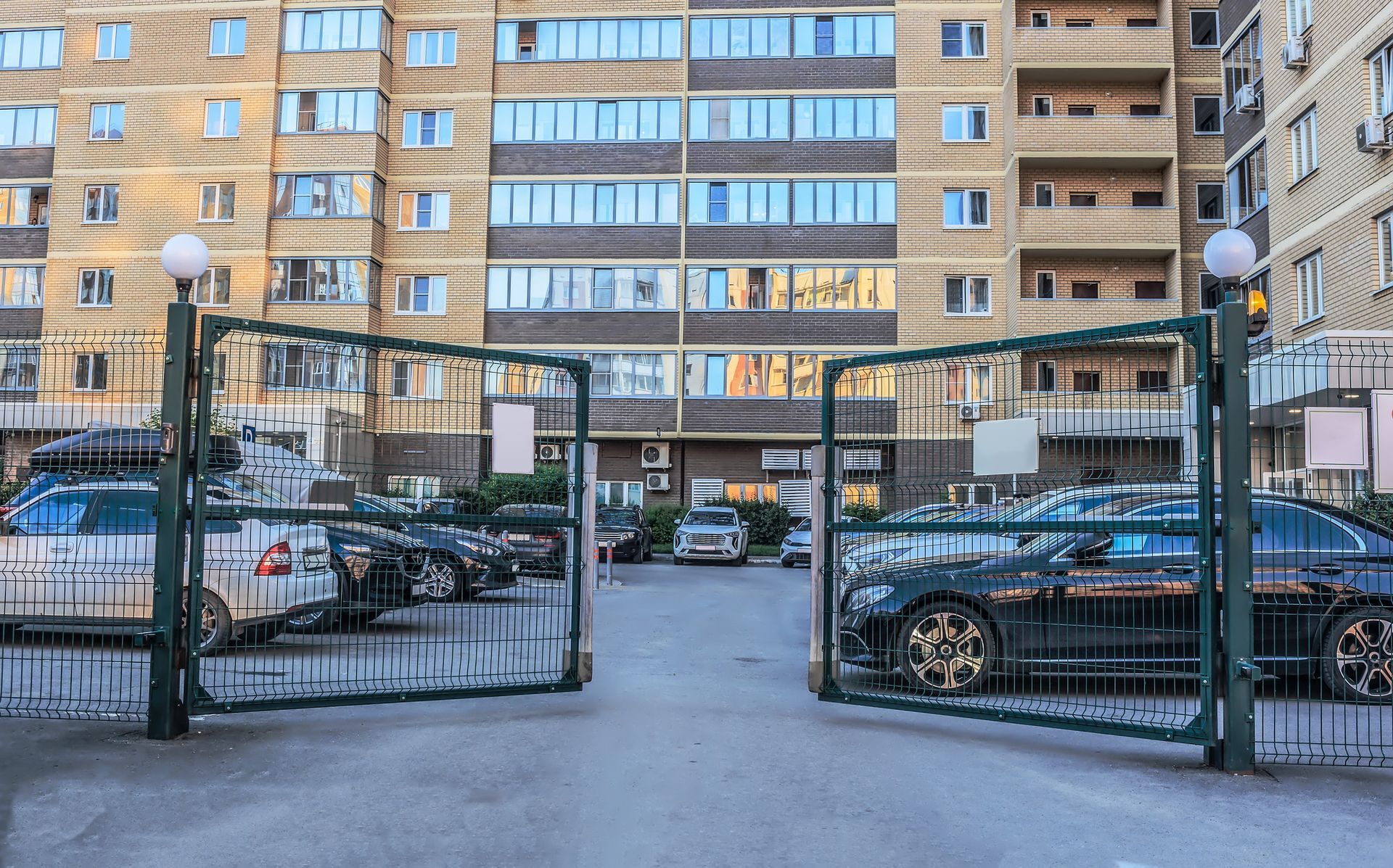 Open metal gate leads to a parking area with cars. Multi-story apartment building in the background.
