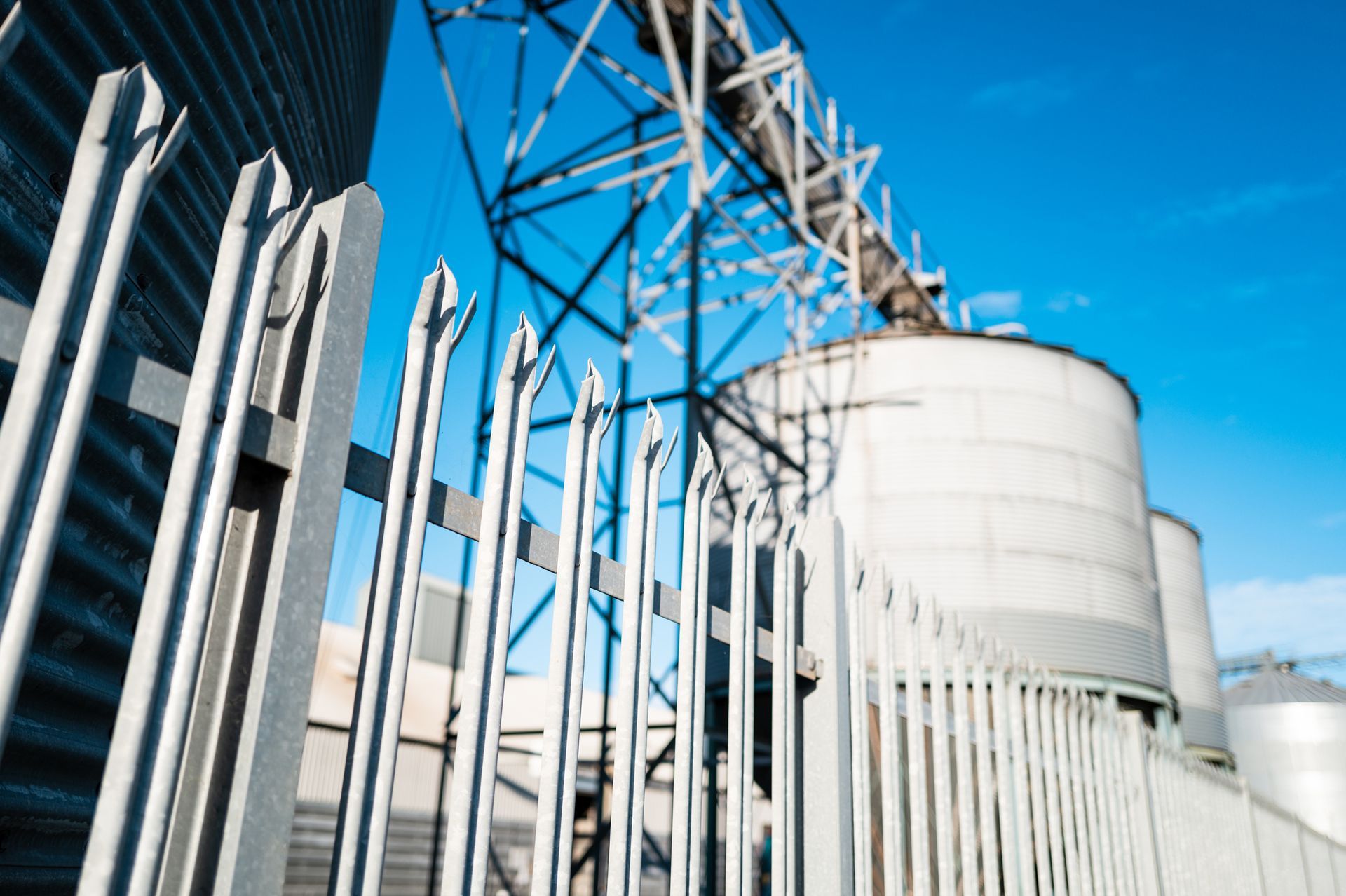 Metal fence in front of grain silos and industrial structure against a blue sky.