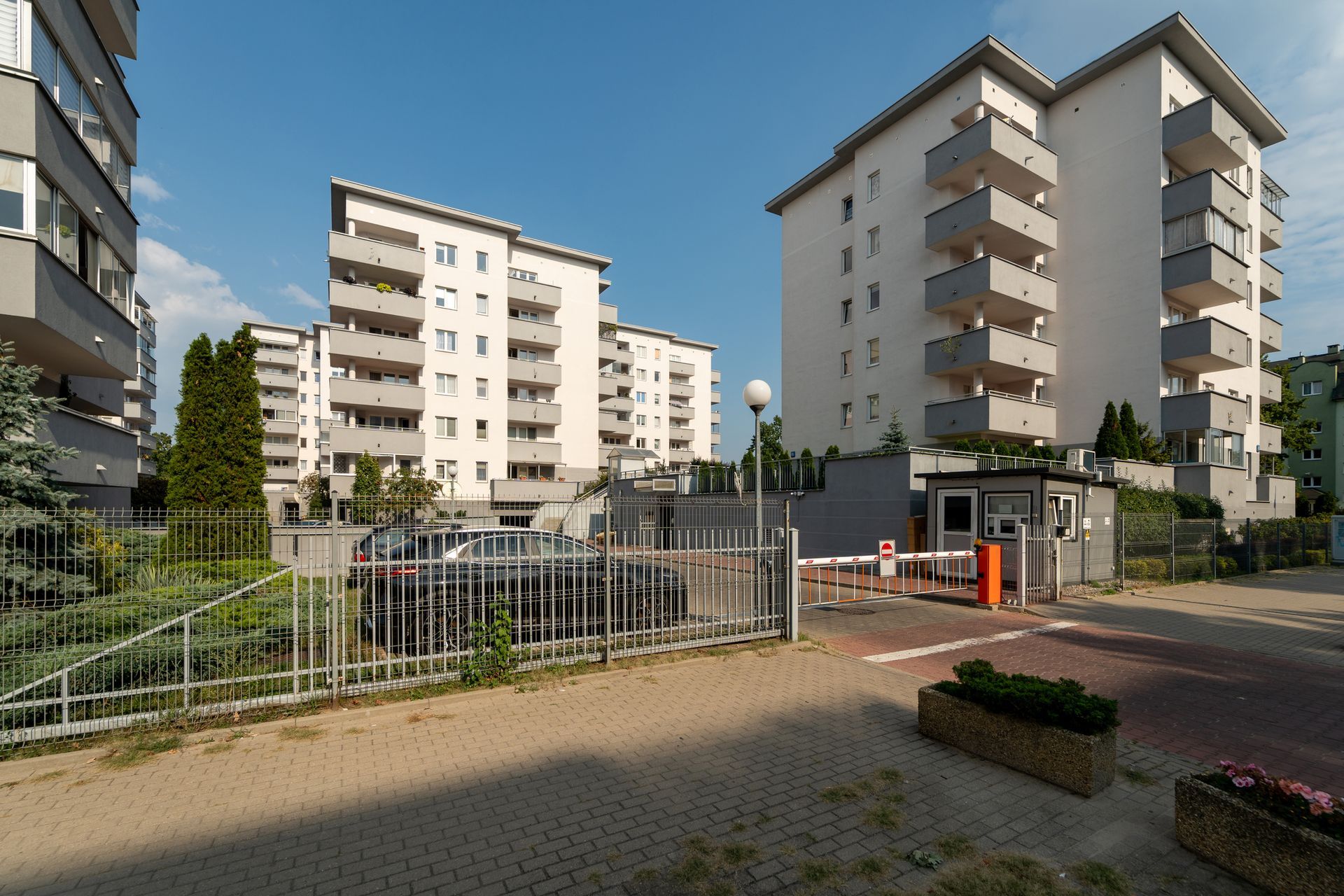 Apartment buildings with balconies, gated entry with cars, blue sky.