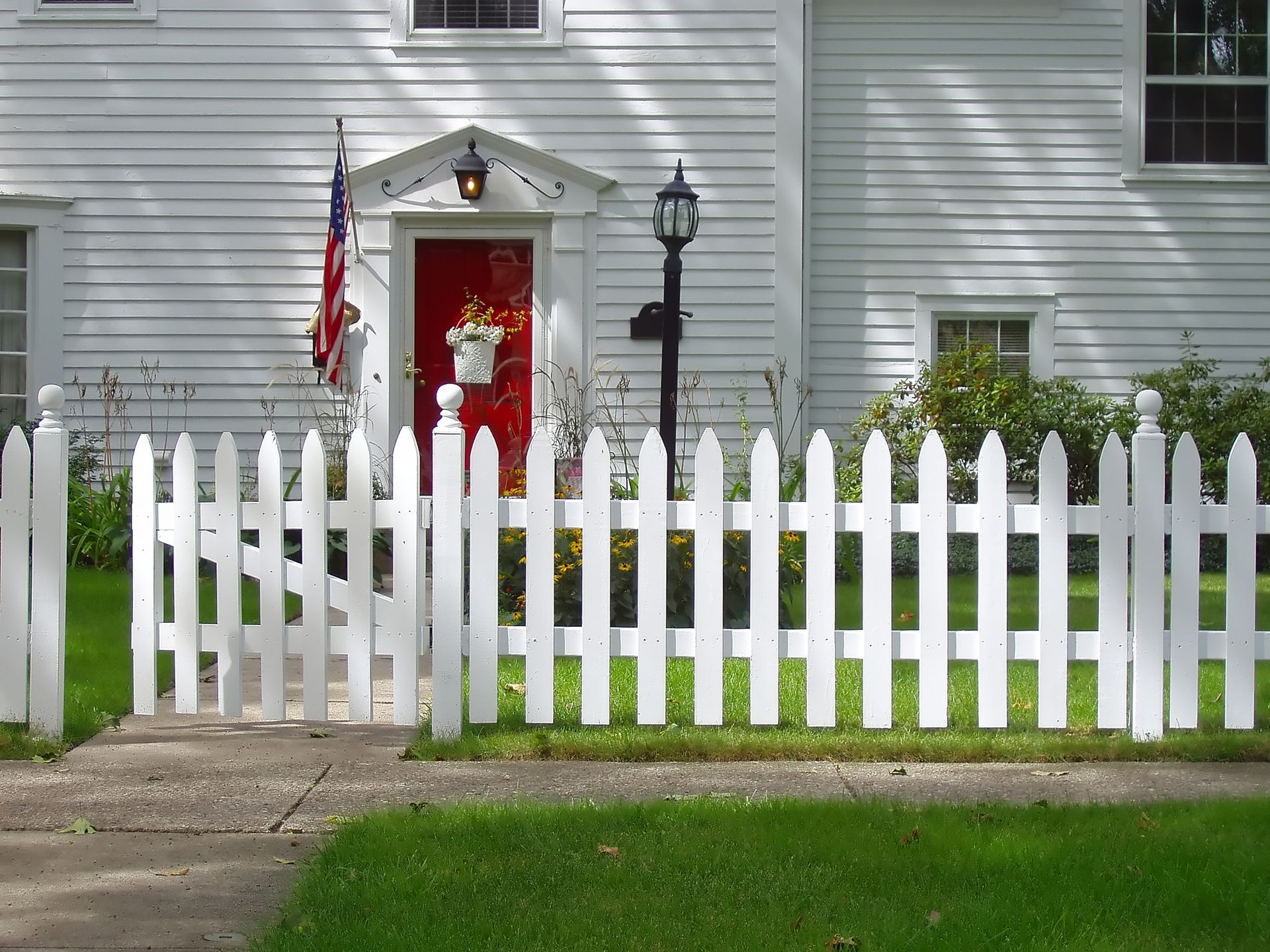 White picket fence in front of a white house with a red door, flowers, and an American flag.