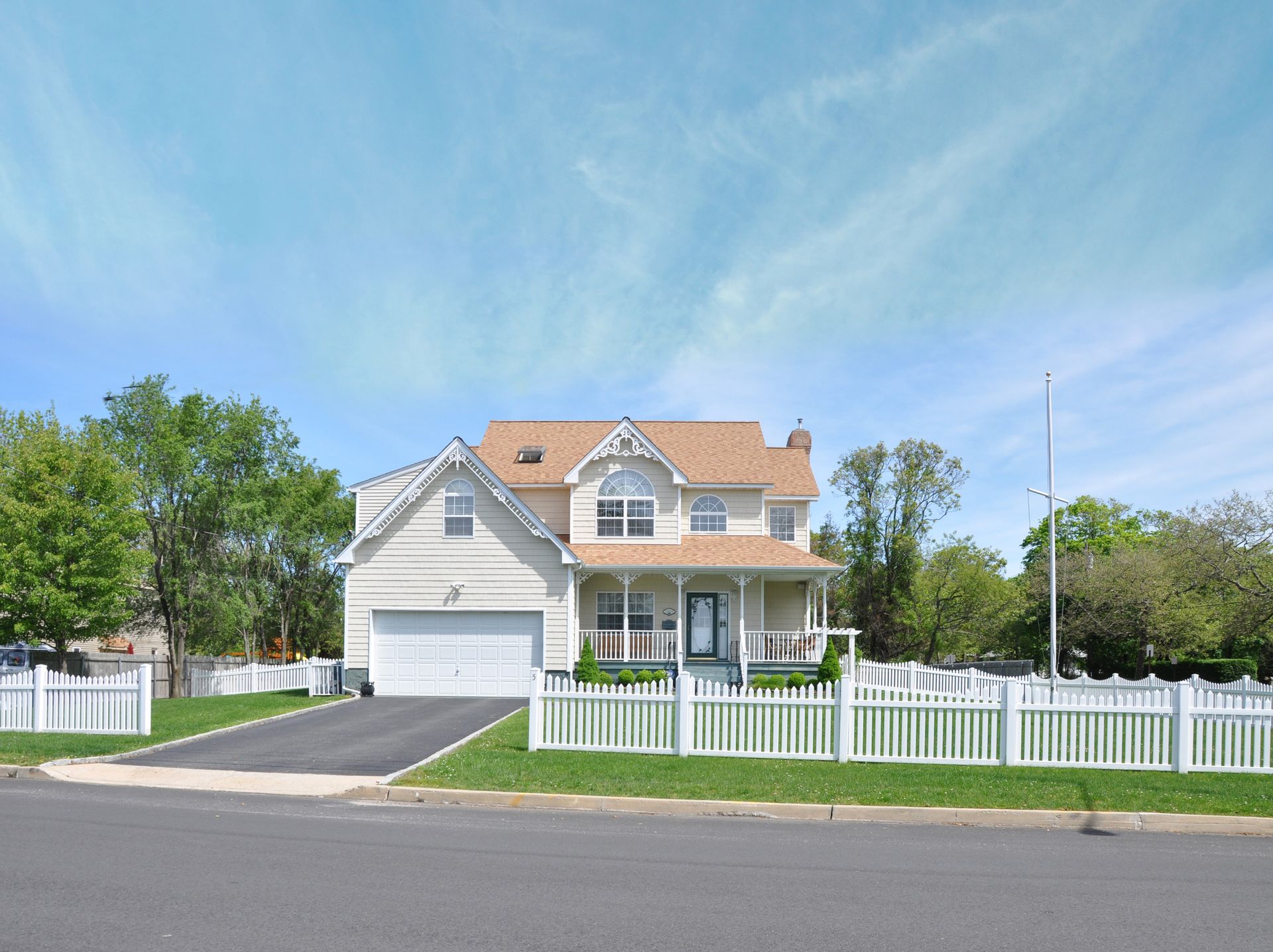Beige house with a white picket fence, driveway, and front porch under a blue sky.
